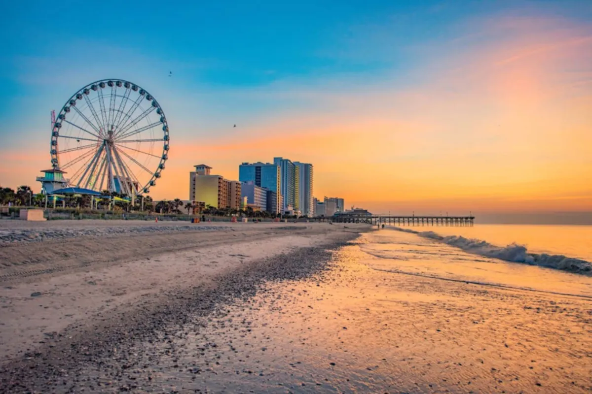 A vibrant exterior shot featuring the Myrtle Beach Ferris wheel, sandy shoreline, blue ocean, and a bustling pier with city skyline in the background. The scene captures families strolling along the beach, the lively boardwalk atmosphere, and the iconic Ferris wheel inviting visitors to experience panoramic views.