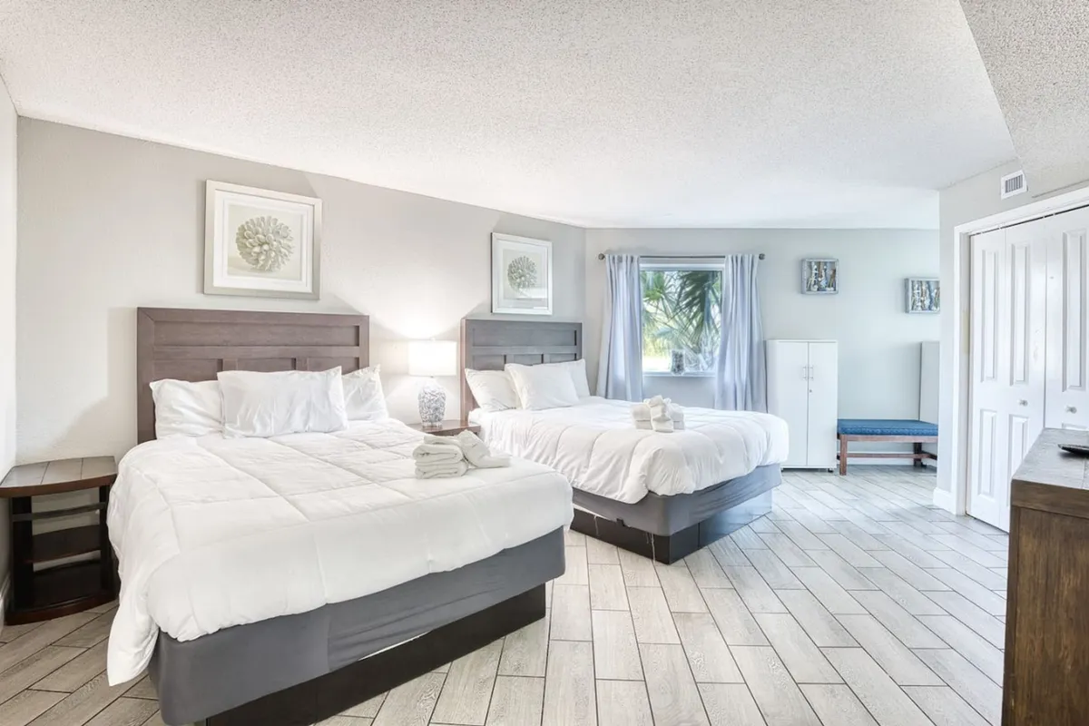 A comfortable bedroom featuring two queen beds with crisp linens, a nightstand with a lamp, and a large window dressed with curtains letting in natural light. The soft ambiance and uncluttered space evoke a restful, welcoming atmosphere ideal for unwinding after a busy day.