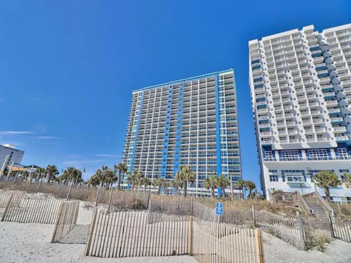 A bright exterior view featuring tall beachfront resort buildings set against a blue sky, with palm trees and sand dunes in the foreground. The scene captures the immediate proximity to the beach and the inviting outdoor atmosphere, with plenty of space for visitors to gather or stroll along the shoreline.