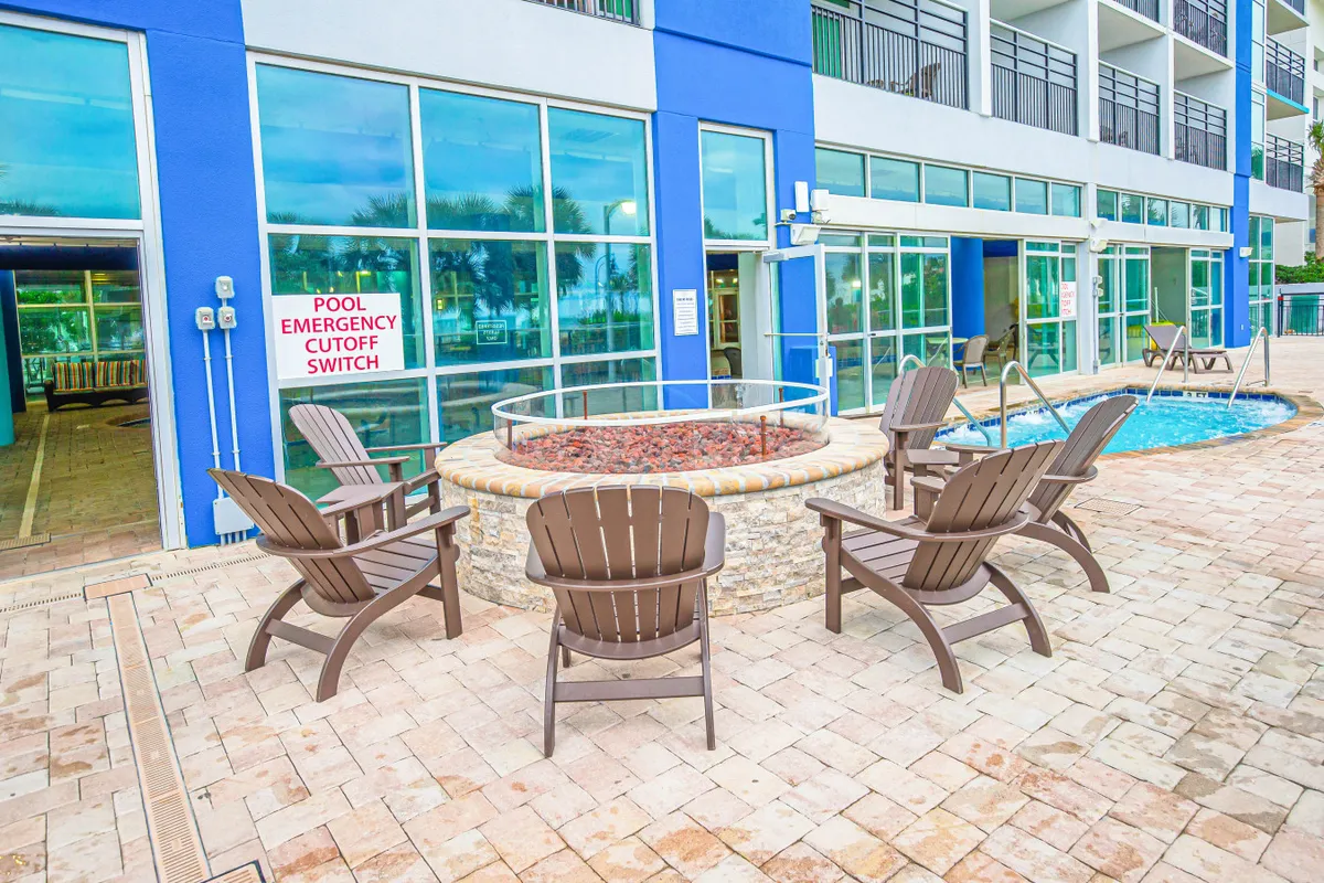 An exterior shot highlighting a fire pit seating area adjacent to the pool, with the building facade in the background. The scene shows guests relaxing by the fire, swimming in the pool, and socializing, capturing the vibrant, resort-style amenities available just steps from the condo.