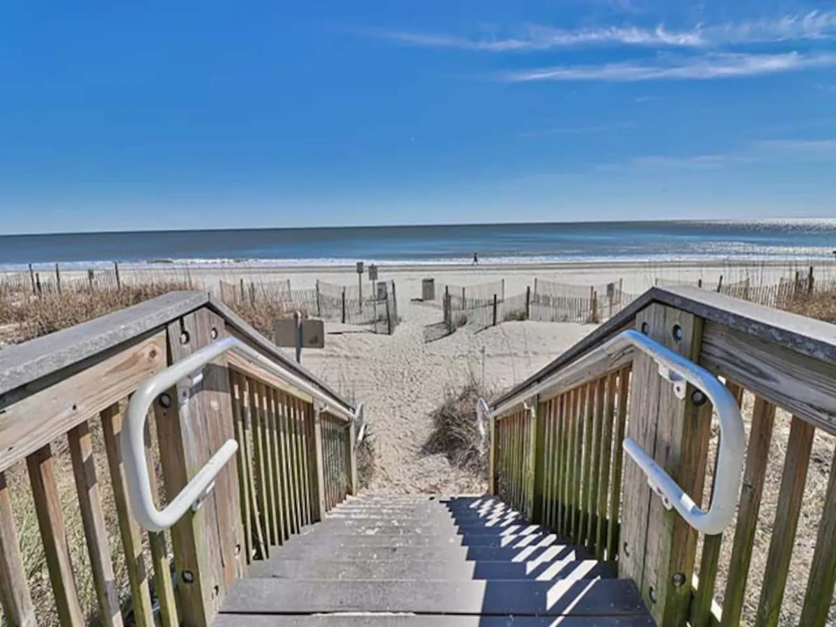 A peaceful boardwalk with weathered wooden steps leading down to the soft sand and expansive ocean, fenced to guide families toward the beach. The image invites viewers to imagine building sandcastles, taking beach walks, and enjoying relaxed family time by the shore.
