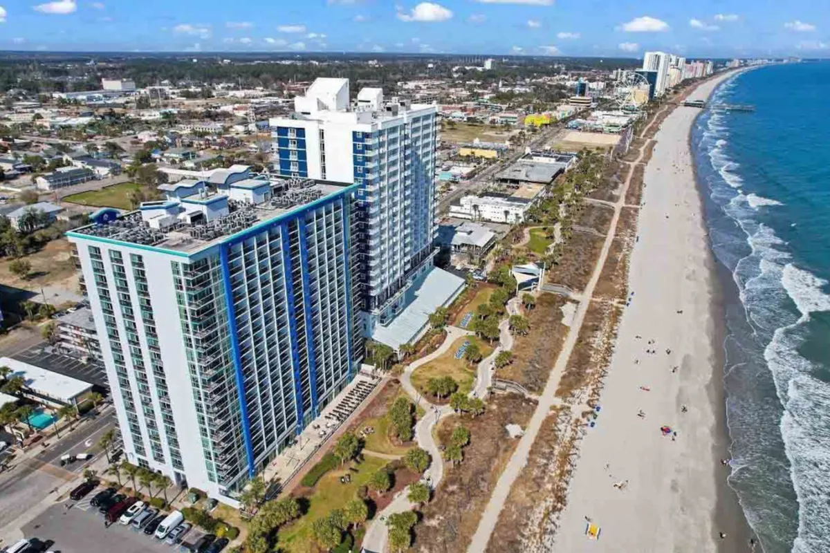 This exterior shot features high-rise beachfront buildings, swaying palm trees, and the ocean waves coming ashore. It highlights the property's central location amidst the beach and activity, with easy access to festivals, events, and local hot spots.