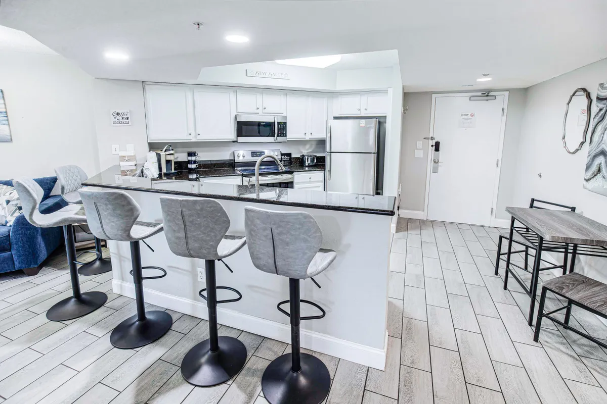 The image showcases a modern kitchen with a spacious island, bar stools, and a dining table set up for meals. The kitchen is open and inviting, featuring updated appliances and plenty of countertop space for cooking and socializing.