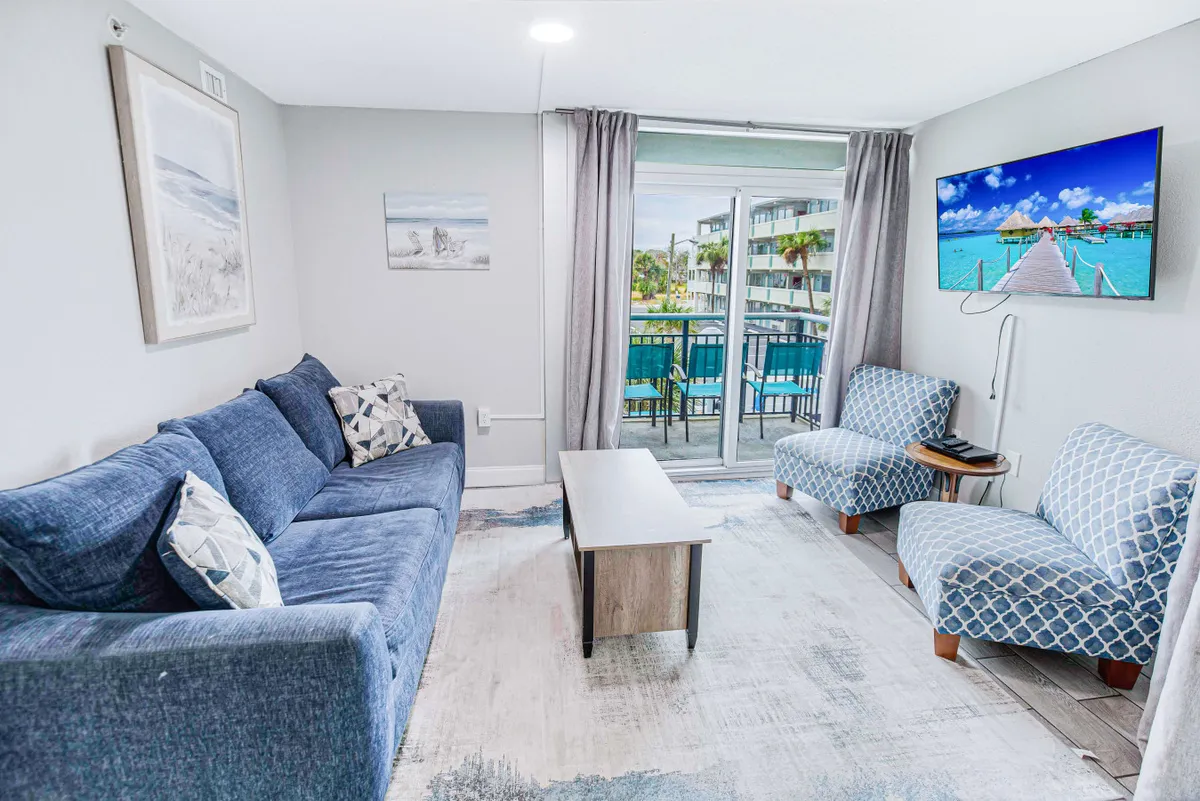 A bright, open living room featuring a comfortable sofa, armchairs, a coffee table, and a TV. Large windows and a sliding glass door provide ample natural light and offer a view of the balcony, seamlessly connecting the interior space to the outdoors.