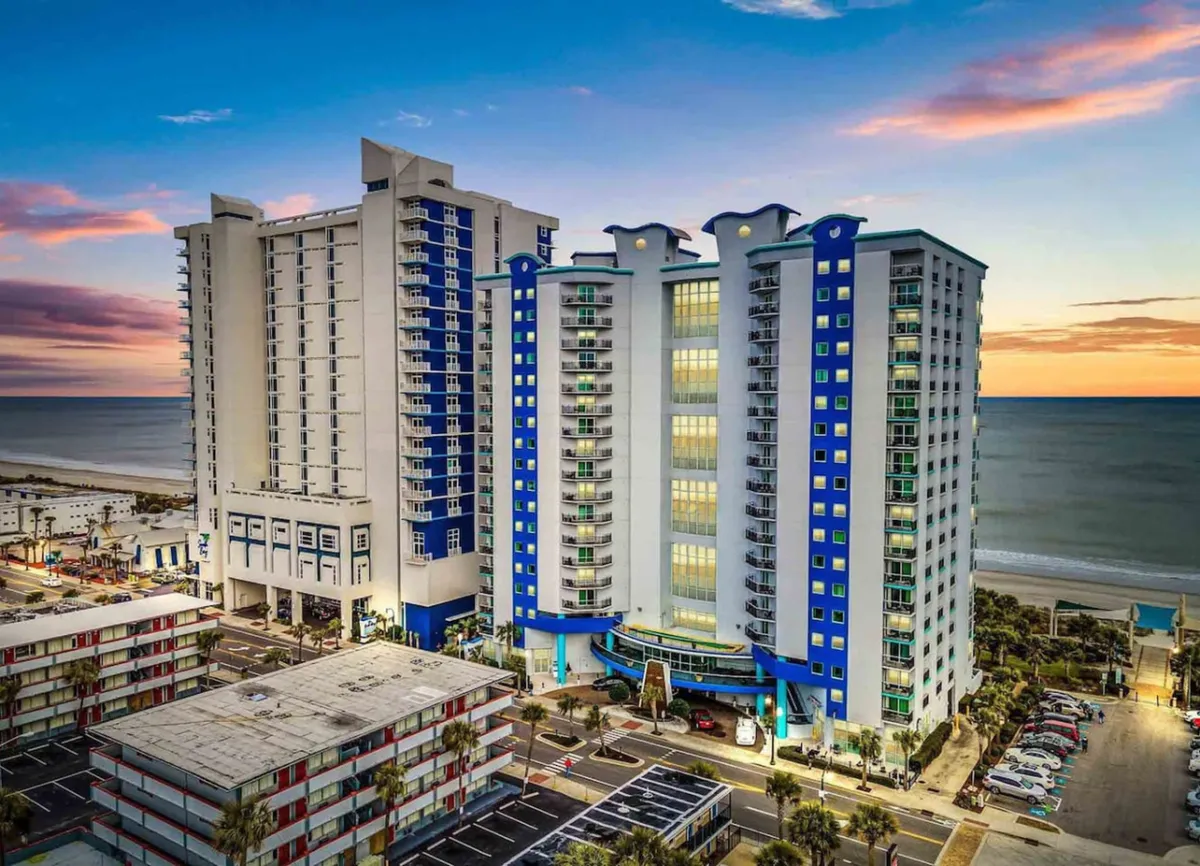 This exterior image captures tall blue and white resort buildings set against a beautiful sunset sky, with an expansive ocean view in the background. The surrounding area hints at easy access to beach activities, shopping, and local dining.