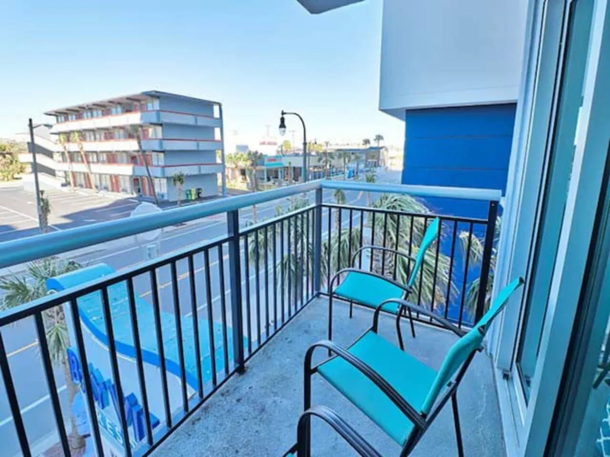 This image shows a balcony with comfortable chairs overlooking a lively street framed by palm trees. The view suggests proximity to local attractions, nightlife, and festival areas. The setting is inviting for relaxing, people-watching, or winding down after an eventful evening.