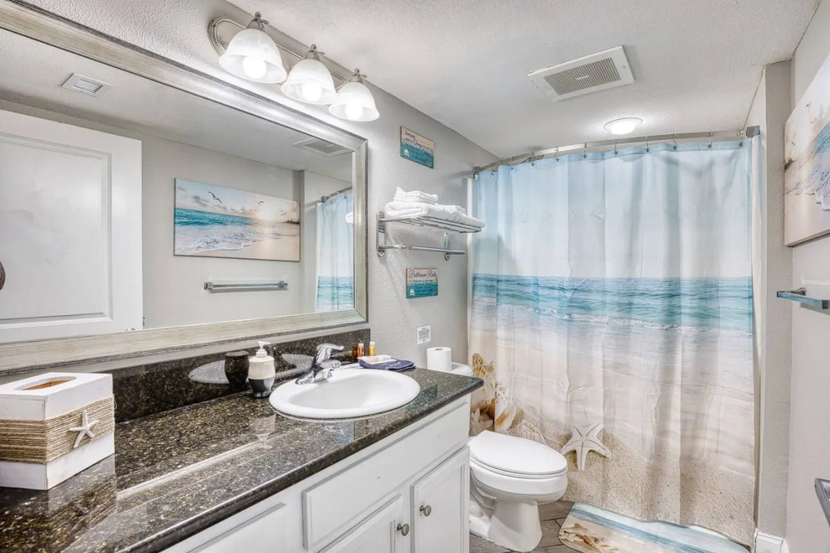 A functional laundry nook with a granite countertop, well-lit mirror, and beach-themed shower curtain in the background. The area feels fresh and well-maintained, suggesting easy access to laundry facilities within the unit.