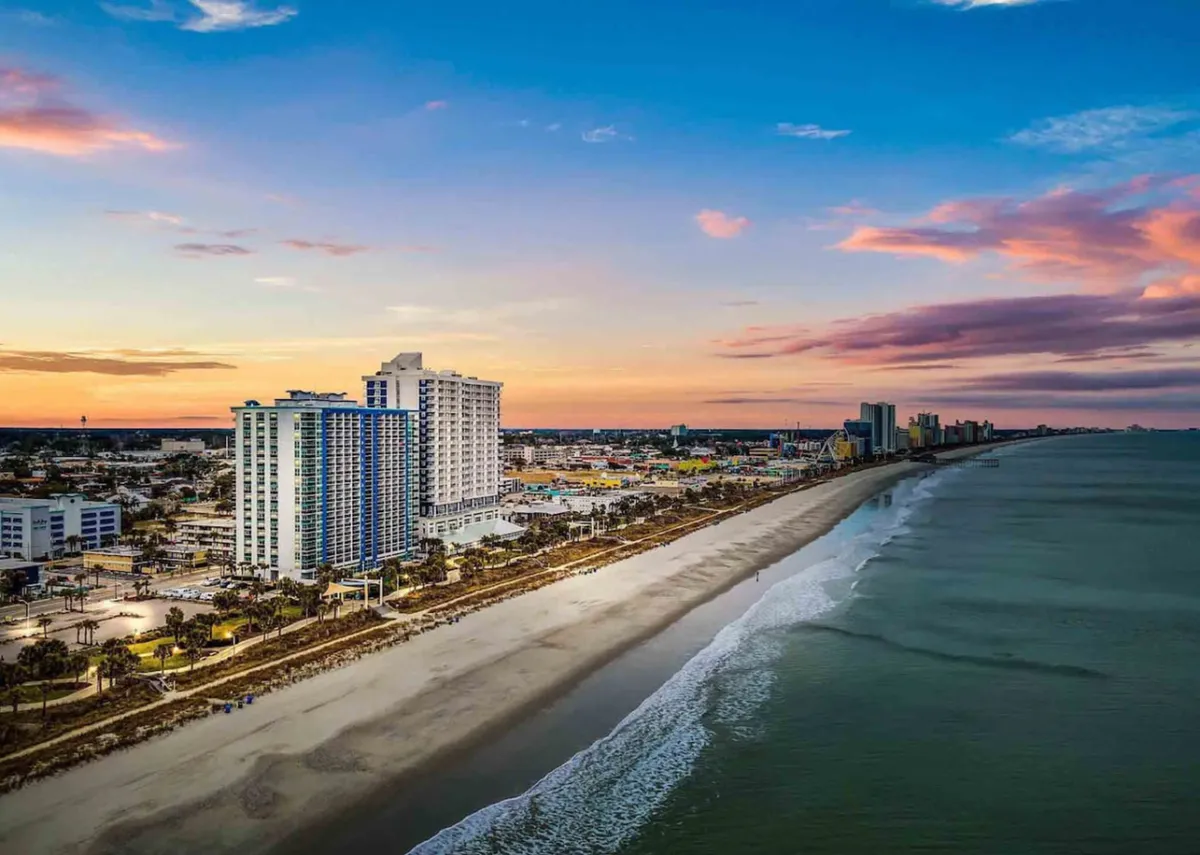 The image showcases a dramatic sunset over Myrtle Beach, with sparkling ocean waves lapping against a sandy shoreline framed by high-rise beachfront condos. The inviting glow of the evening sky creates a warm ambiance, while bustling beachfront activity hints at both relaxation and excitement. The scene highlights direct beach access, breathtaking ocean views, and proximity to vibrant local attractions.