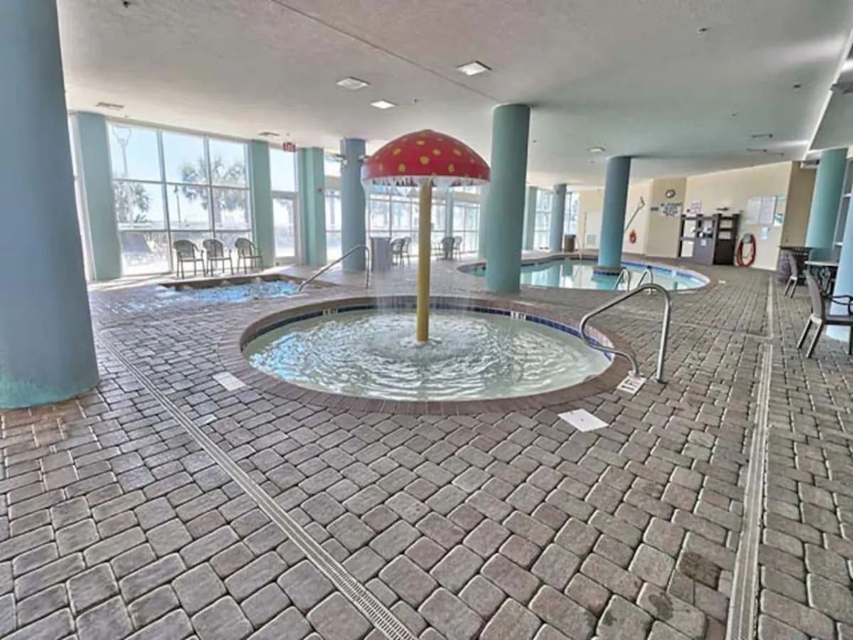 An indoor space featuring cheerful water play with a mushroom-shaped fountain and multiple hot tubs. Adults are seen unwinding in the hot tubs, while children enjoy the playful water feature. Expansive windows allow natural light to stream in, connecting the interior to the outside.